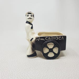 May include: A ceramic figurine of a man pushing a dark brown cart with a cream-colored wheel. The cart has the words "Rio Carioca" on the side. The man is wearing a white outfit with a black tie and hat.