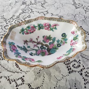 May include: A white, scalloped-edge dish with a floral design featuring pink flowers and green leaves. The dish has a gold rim and a gray border. The floral pattern is centered on the dish.