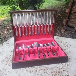 May include: A wooden flatware storage chest with a hinged lid, featuring a bright red interior lining. The chest contains a set of silver-toned metal knives arranged in the lid and a collection of forks and spoons organized in the base.