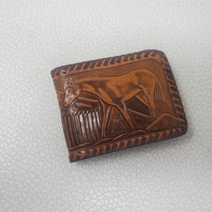 May include: A brown leather bi-fold wallet featuring a detailed embossed design of a horse standing in a field with a fence. The edges of the wallet are decorated with a repeating diagonal stitched pattern, creating a textured border around the image.