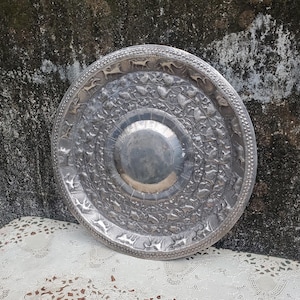 May include: A silver-colored, round metal tray with intricate embossed designs. The tray features a central, slightly recessed area and detailed patterns of animals and floral motifs around the rim. The tray is displayed on a white lace tablecloth.