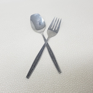 Pode incluir: Uma colher e um garfo de aço inoxidável estão cruzados sobre um fundo texturizado esbranquiçado. Os talheres têm um padrão floral decorativo nas alças. A colher tem formato oval e o garfo tem quatro dentes.