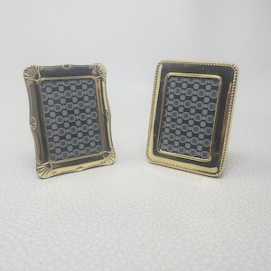 Pode incluir: Dois pequenos porta-retratos retangulares com bordas de metal dourado polido. A moldura da esquerda apresenta um design de borda recortada, enquanto a da direita tem uma borda com contas. Ambas contêm um inserto preto com padrão branco.