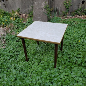 May include: A small, square table with a light-colored, marble-like top and dark brown legs. The table has a gold-colored trim around the top. The table is set outdoors on a bed of green foliage.