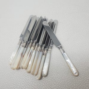 May include: A set of nine silver-plated dinner knives featuring polished metal blades and smooth, iridescent mother-of-pearl handles. Each knife has a decorative silver collar connecting the blade to the handle, arranged in a fan-like cluster.