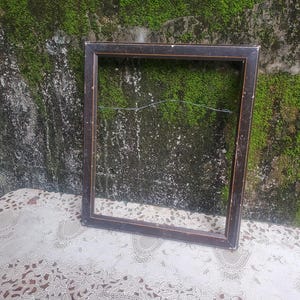 Pode incluir: Uma moldura retangular com acabamento em castanho escuro e sinais visíveis de desgaste, incluindo riscos leves e arranhões ao longo das bordas. A moldura apresenta um detalhe de borda interna fina e clara e um fio de metal na parte traseira.