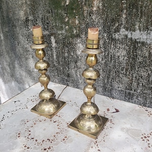 May include: Two antique brass table lamps with ornate detailing. Each lamp has a square base, a decorative stem, and a cylindrical top with a light bulb. The lamps are placed on a white lace tablecloth.