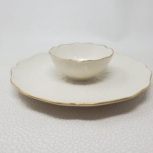 May include: A white ceramic serving set consisting of a large, round, scalloped-edge plate and a smaller, matching scalloped bowl centered on top. Both pieces feature a delicate, embossed sunburst pattern and a thin, metallic gold rim.
