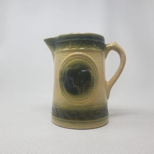 May include: A ceramic jug with a cream-coloured body and a dark green rim and base. The jug features a raised design and a dark green emblem on the front. It has a curved handle and a spout.