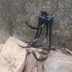 May include: A black metal garden tool with curved tines and a decorative top. The tool is designed for digging or cultivating soil. The image shows the tool resting on a wooden surface.