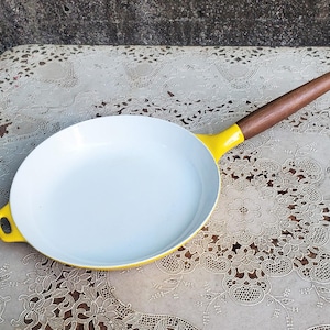 May include: A round frying pan with a bright yellow exterior and a smooth white interior. The pan features a long, straight brown wooden handle and a small, integrated yellow loop handle on the opposite side for easy lifting and storage.