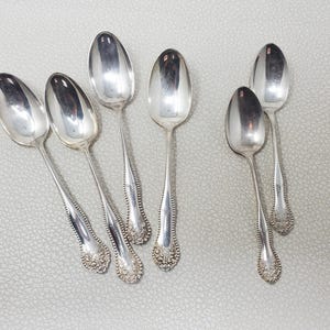 May include: Six silver spoons with oval bowls and ornate, slender handles. The spoons are arranged on a light grey surface. The handles feature a floral design at the base, and the spoons reflect light.