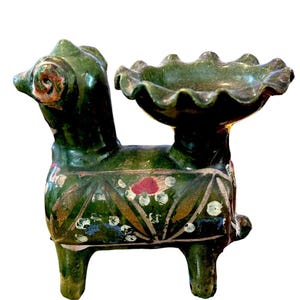 May include: A green ceramic animal-shaped candle holder with a ruffled bowl. The candle holder is decorated with floral patterns in red, white, and gold. The animal has a long neck and a detailed face.