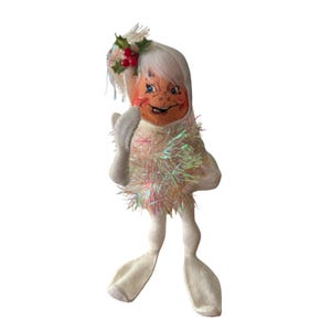 May include: A vintage Christmas doll with a smiling face, white hair, and a holly berry accent. The doll is wearing a white outfit with iridescent tinsel trim and white gloves and shoes. A whimsical holiday decoration.