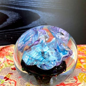 May include: A clear glass paperweight with a swirling design of blue, purple, and orange colors. The interior features a dark base, and the overall effect is reminiscent of an abstract landscape. The paperweight is spherical in shape.