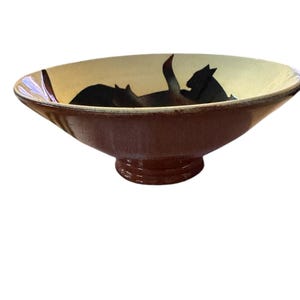 May include: A ceramic bowl with a wide, shallow design. The exterior is a deep brown, while the interior features a cream-colored background with black cat silhouettes. The bowl has a pedestal base.