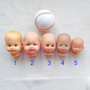 Puede incluir: Cinco cabezas de muñecas vintage de varios tamaños y tonos de piel están dispuestas en una fila, con una pelota de béisbol encima. Las cabezas de las muñecas tienen rasgos faciales pintados, incluidos ojos y bocas. La pelota de béisbol es blanca con costuras rojas.