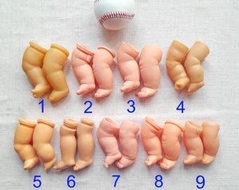 May include: A collection of nine pairs of doll legs in varying shades of peach and tan, arranged with a baseball. Each pair is numbered in blue. The baseball is white with red stitching.