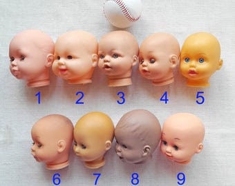 May include: Nine vintage doll heads in various skin tones, numbered 1-9, with painted facial features. A white baseball with red stitching is positioned above the doll heads. The doll heads are made of plastic and are a collectible item.