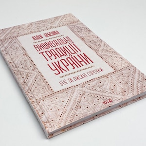 Puede incluir: Un libro de tapa dura titulado "Tradiciones de bordado ucranianas" en texto rojo sobre fondo blanco. La portada del libro presenta un diseño bordado en tonos beige y marrón. El libro está ligeramente inclinado.