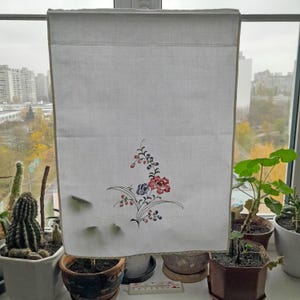 Könnte beinhalten: Weißer Leinenvorhang mit einem floralen Stickmuster mit rosa, blauen und grünen Blumen. Der Vorhang hängt an einem Fenster mit Blick auf eine Skyline und Bäume.