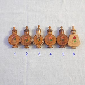 以下が含まれることがあります： 精巧なデザインの小さな木製香水瓶が6つ。各ボトルには、バラの絵と「ブルガリア」の文字が描かれています。ボトルは様々な形をしており、装飾的なストッパーが付いています。