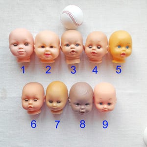 May include: Nine doll heads with varying skin tones and facial expressions are arranged in a row. A white baseball sits above the doll heads. Each doll head has a unique number from 1 to 9.