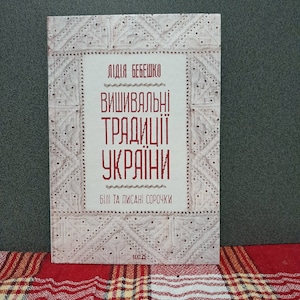 Op de afbeelding: Een boek getiteld "Oekraïense borduurtradities" van Lidiia Bebeshko. De omslag heeft een wit geborduurd ontwerp met de titel in rode tekst. Het boek ligt op een rood-wit geruit oppervlak.