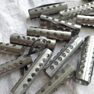 Peut inclure: Gros plan sur une collection de bigoudis en métal argenté. Chaque bigoudi est cylindrique, percé de trous réguliers et doté d'une attache métallique. Le fond est un tissu blanc texturé.