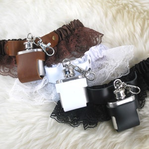 May include: A selection of hip flasks in brown, white, and black leather cases, each attached to a garter with lace trim. The flasks have a silver finish and are displayed on a fluffy, white surface.