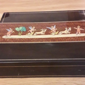 May include: A black wooden box with a decorative inlay of cherubs riding goats. The inlay is on a brown background with green trees.