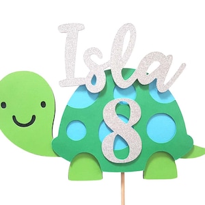 May include: Green and blue turtle cake topper with the name Isla and the number 8 in silver glitter.