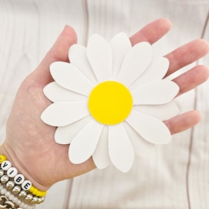 May include: A white paper daisy with a yellow center. The daisy is being held in a person's hand.