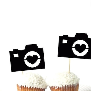 May include: Two black cupcake toppers shaped like cameras with a heart in the lens. The toppers are on toothpicks and are ready to be placed on cupcakes.