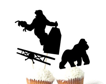 May include: Black silhouette cupcake toppers featuring a gorilla climbing a building, a biplane, and a gorilla walking. The toppers are placed on cupcakes with white frosting. Perfect for a themed party.