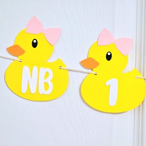 May include: Yellow rubber ducky banner with pink bows and the letters "NB" and the number "1" on each duck. The banner is strung together with white string.