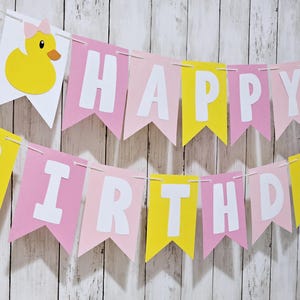 May include: A festive banner with the words "HAPPY BIRTHDAY" in white letters on pink and yellow pennants. The banner is decorated with yellow duck cutouts, perfect for a birthday celebration.