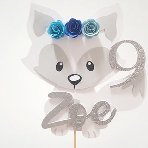 May include: White fox cake topper with a blue flower crown and the name "Zoe" in silver glitter. The fox is holding a silver glitter number 9.