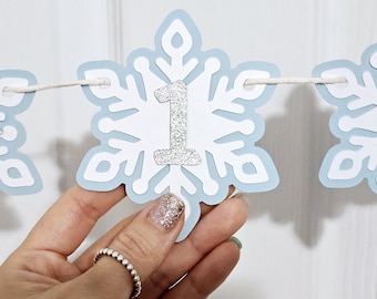 May include: A blue and white snowflake banner with a silver glitter number one. The banner is strung with white twine.