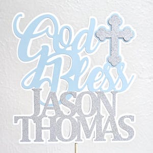 May include: A light blue and silver glitter cake topper with the words "God Bless" and the name "Jason Thomas". The topper includes a silver glitter cross and is attached to a wooden stick.