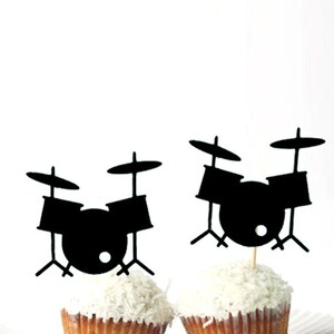 May include: Two black cupcake toppers shaped like drum sets. Each topper features a bass drum, two smaller drums, and two cymbals.