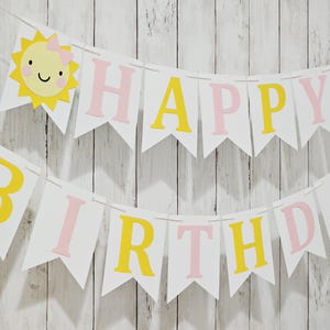 May include: A white banner with the words "HAPPY BIRTHDAY" in pink and yellow letters. The banner is decorated with smiling sun cutouts. The banner is strung on a white string and is against a white wooden background.