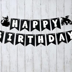 May include: Black and white banner with the words "HAPPY BIRTHDAY" in white block letters. The banner is strung between two black dragon silhouettes, against a white wooden background. A festive decoration for a birthday celebration.