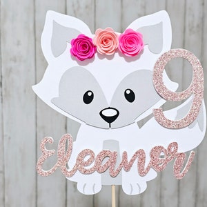 May include: A white fox cake topper with a pink glitter number 9, the name "Eleanor" in pink glitter, and a pink flower crown.