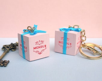May include: Two pink gift box keychains with blue ribbon and gold keychains. The boxes have the text "Mendl's" printed on them.