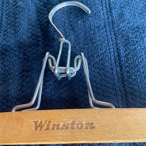 May include: A wooden clothes hanger with a metal hook and clips. The word "Winston" is engraved on the wooden bar. The hanger is designed for clothing storage.