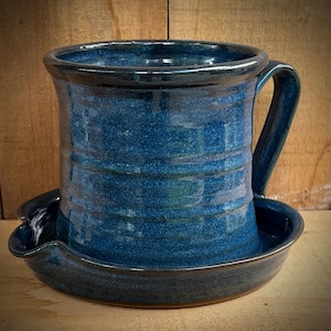 May include: A deep blue ceramic mug with a glossy finish and horizontal ribbed texture, featuring a sturdy handle and a matching circular saucer base with a small integrated spout on the side, showcasing a rich, mottled cobalt glaze throughout.