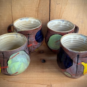 May include: Four ceramic mugs with a rounded shape and ribbed interior texture. Each mug features a reddish-brown base color decorated with abstract, hand-painted shapes in yellow, light green, and dark blue, accented by thin black and red lines.
