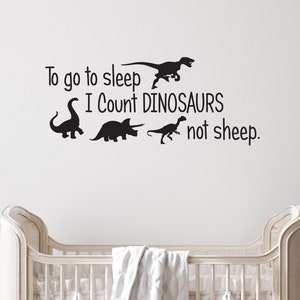 May include: A white crib with a white blanket in it. The wall behind the crib has a black vinyl decal that says "To go to sleep I count dinosaurs not sheep." There are four dinosaur silhouettes in the decal.