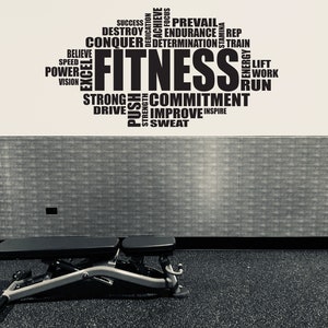 Puede incluir: Imagen en blanco y negro de un gimnasio. Un banco de pesas se encuentra sobre un suelo de goma negro frente a una pared con la palabra "FITNESS" rodeada de palabras motivadoras como "ÉXITO" y "COMPROMISO".